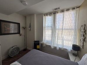 A bedroom with a bed, a standing fan, a heater, and a window with patterned curtains.