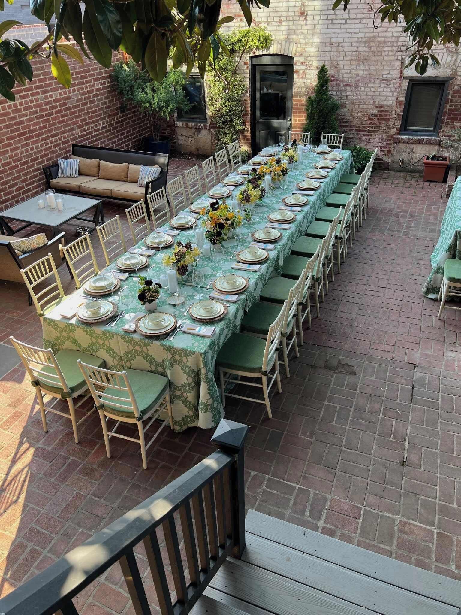 A long dining table set with chairs, a sofa, and a small table with drinks, all outdoors with a brick wall and greenery in the background.