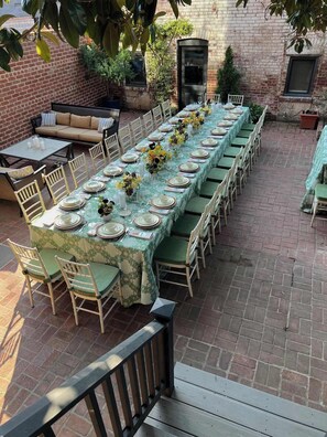 A long dining table set with chairs, a sofa, and a small table with drinks, all outdoors with a brick wall and greenery in the background. - The Manor on Vermont, Historic DC Luxury Retreat (Washington)