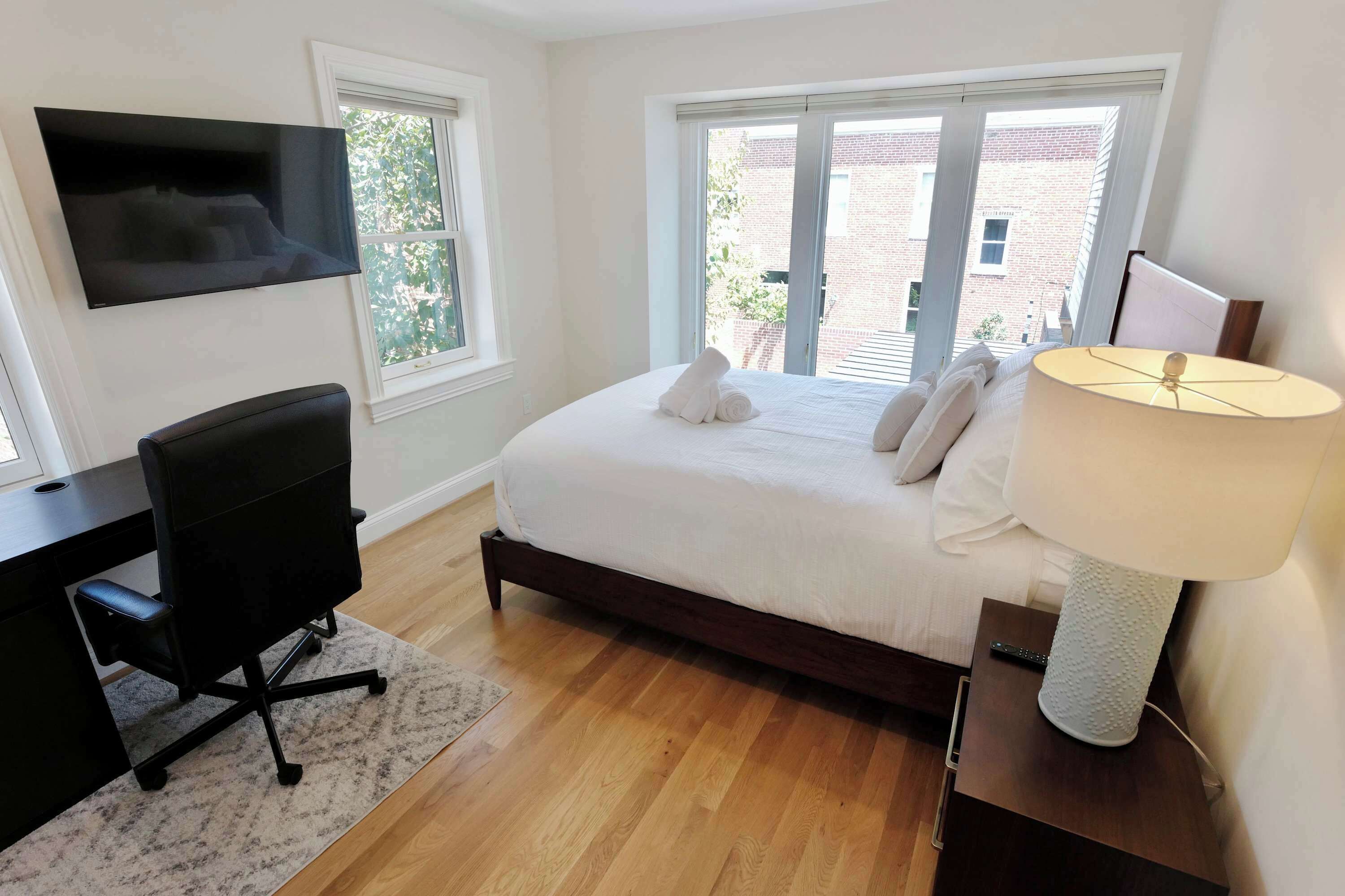A bedroom with a bed, a desk, a chair, a TV, and a window with a view of a brick building.