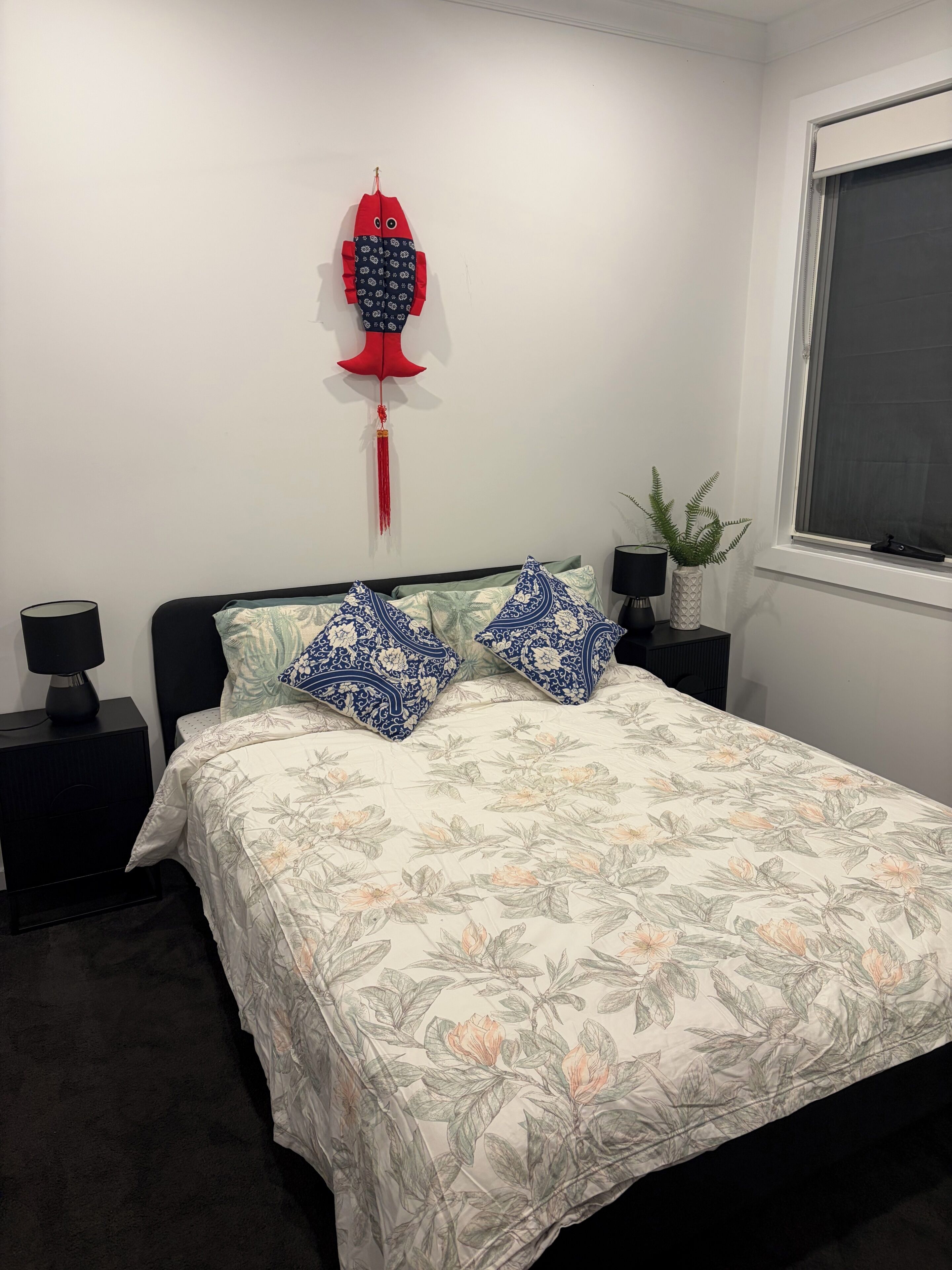 A bedroom with a bed, two nightstands, a red decorative item on the wall, a potted plant, and a window.