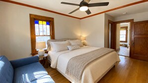 Un dormitorio con cama, ventilador de techo, ventana con vitrales, mesita de noche con lámpara y puerta que da a otra habitación.