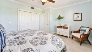 Un dormitorio con una cama, ventilador de techo, cómoda, una silla y un cuadro en la pared.