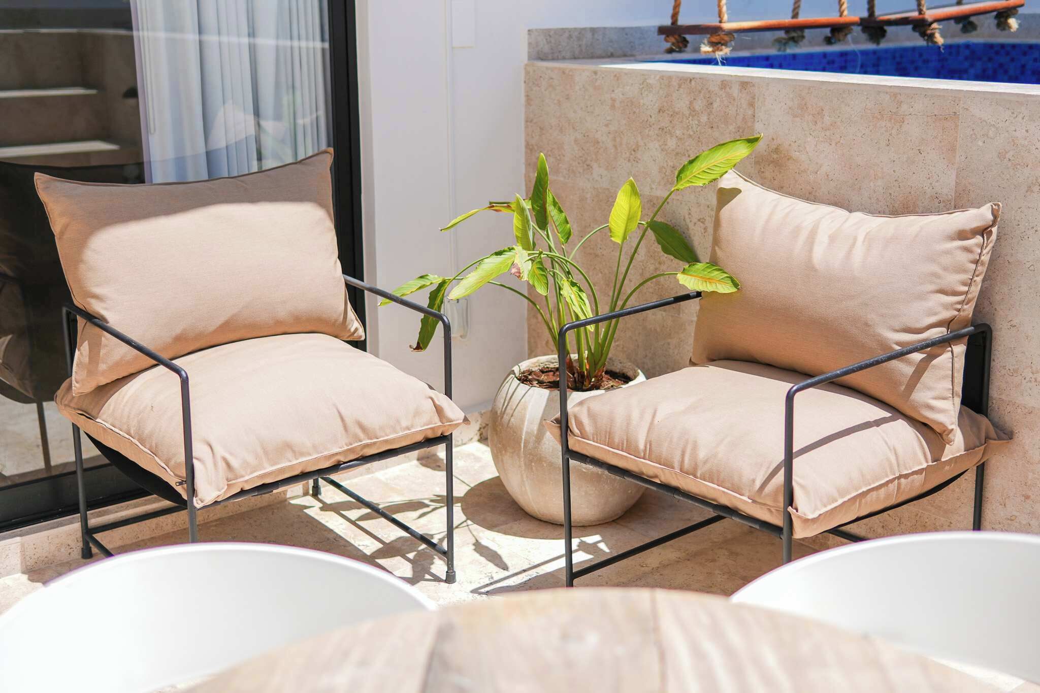 Dos sillones color beige con estructura metálica, una planta en maceta y una mesa blanca en un balcón.