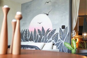 A room with a large wall mural of birds flying over a sunset, a sofa with cushions, and a plant on the windowsill. - Modern 2BR Retreat in Parques Zama w/ Private Pool (Tulum)