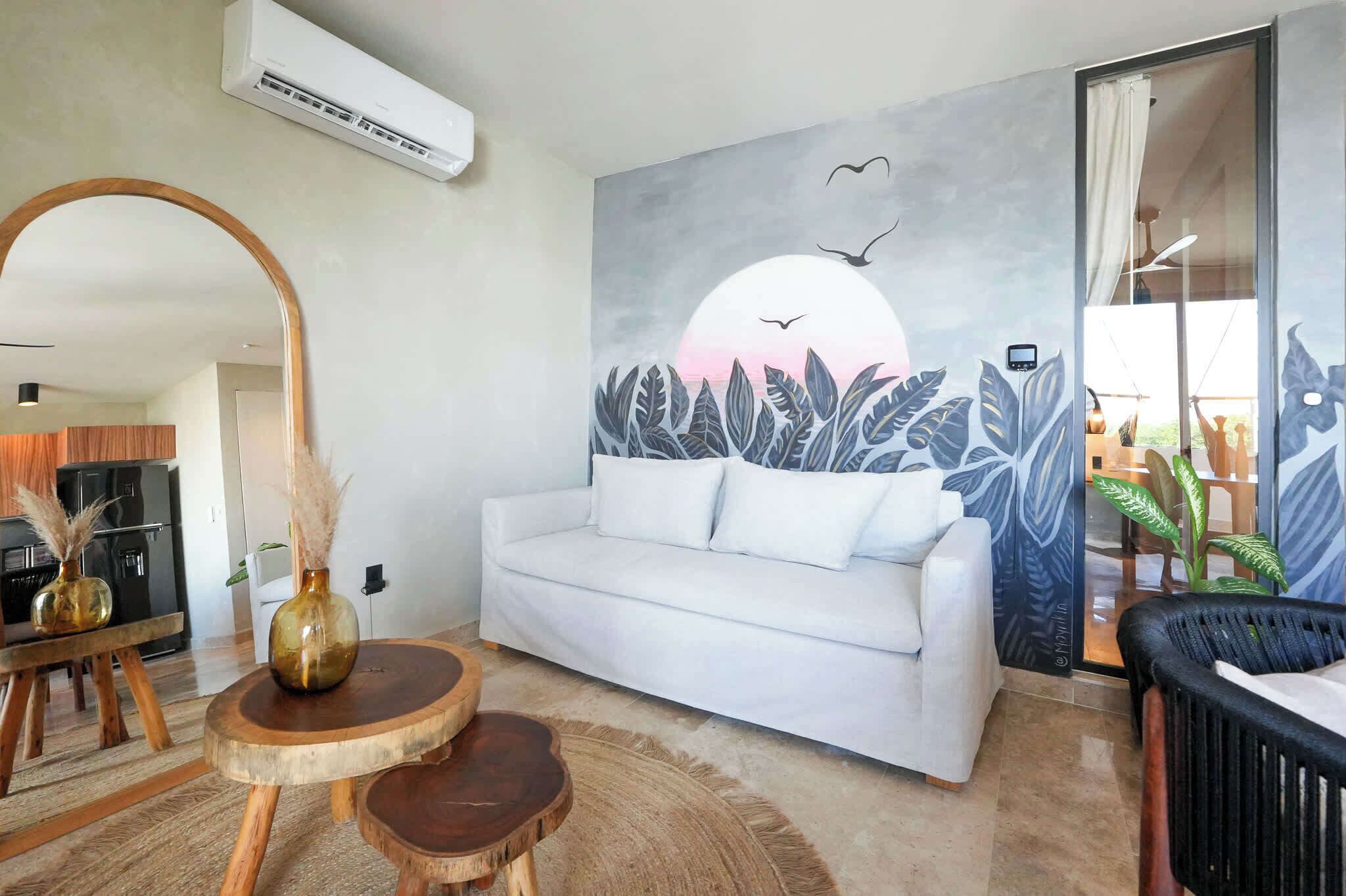 A modern living room with a white sofa, a round wooden coffee table, and a wall mural depicting a sunset with birds.