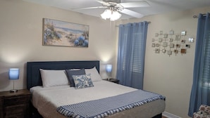 A bedroom with a bed, two bedside lamps, a ceiling fan, a painting, and a wall-mounted mirror.