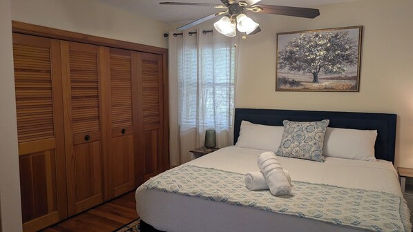 A bedroom with a bed, a ceiling fan, a nightstand, a lamp, a painting, and a wooden wardrobe.