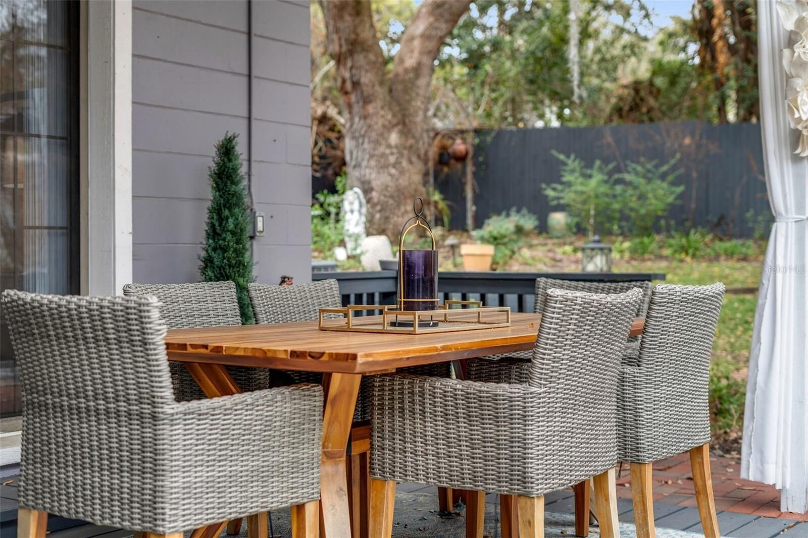 A patio with a wooden table and wicker chairs, surrounded by greenery.
