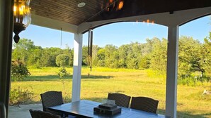 Um pátio externo com mesa e cadeiras, um ventilador de teto e vista para um campo verde.