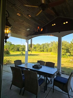 Eine Terrasse im Freien mit Tisch und Stühlen, einem Deckenventilator und Blick auf ein grünes Feld.