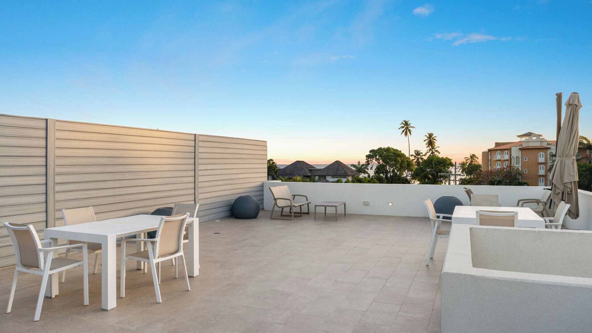 Un patio al aire libre con mobiliario blanco, un muro bajo y vistas a un resort con palmeras y un atardecer.