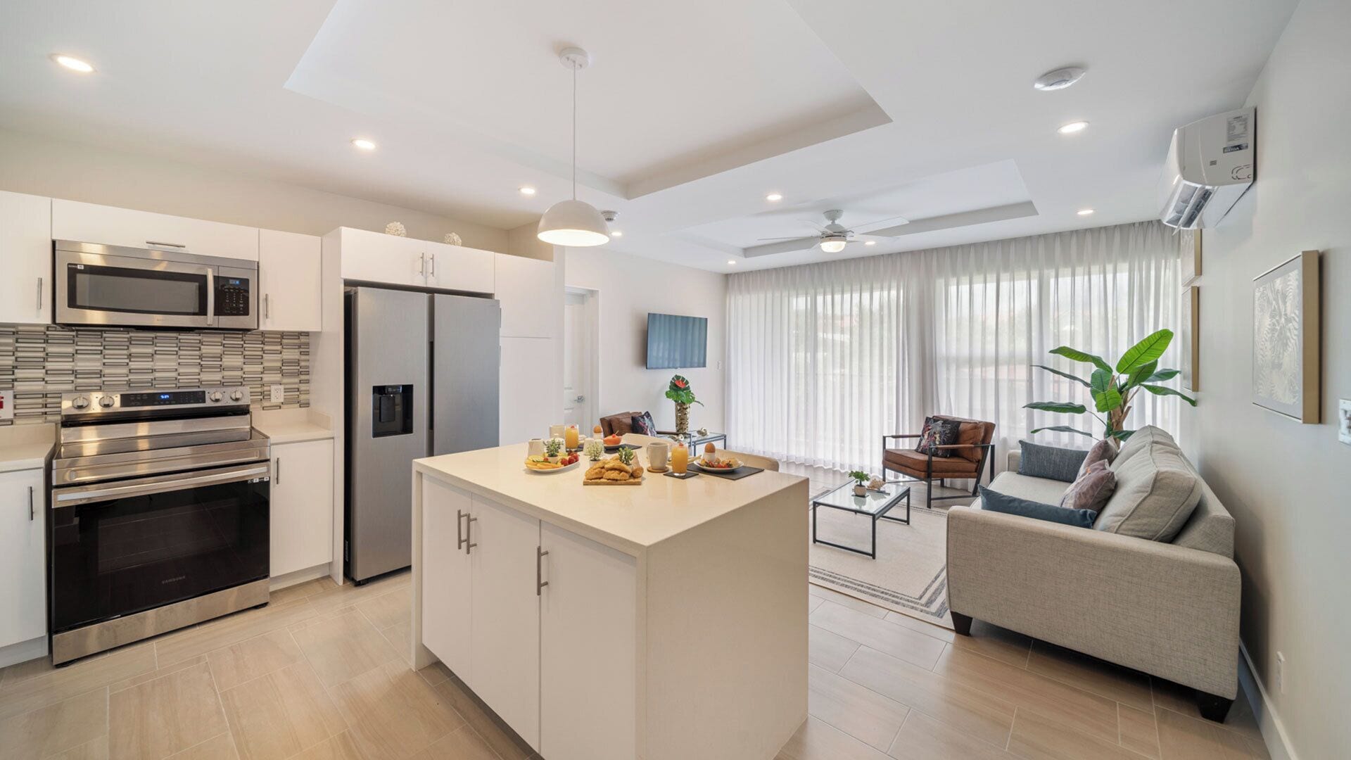 A modern kitchen with stainless steel appliances, a central island with breakfast items, and a living area with a sofa and a coffee table.