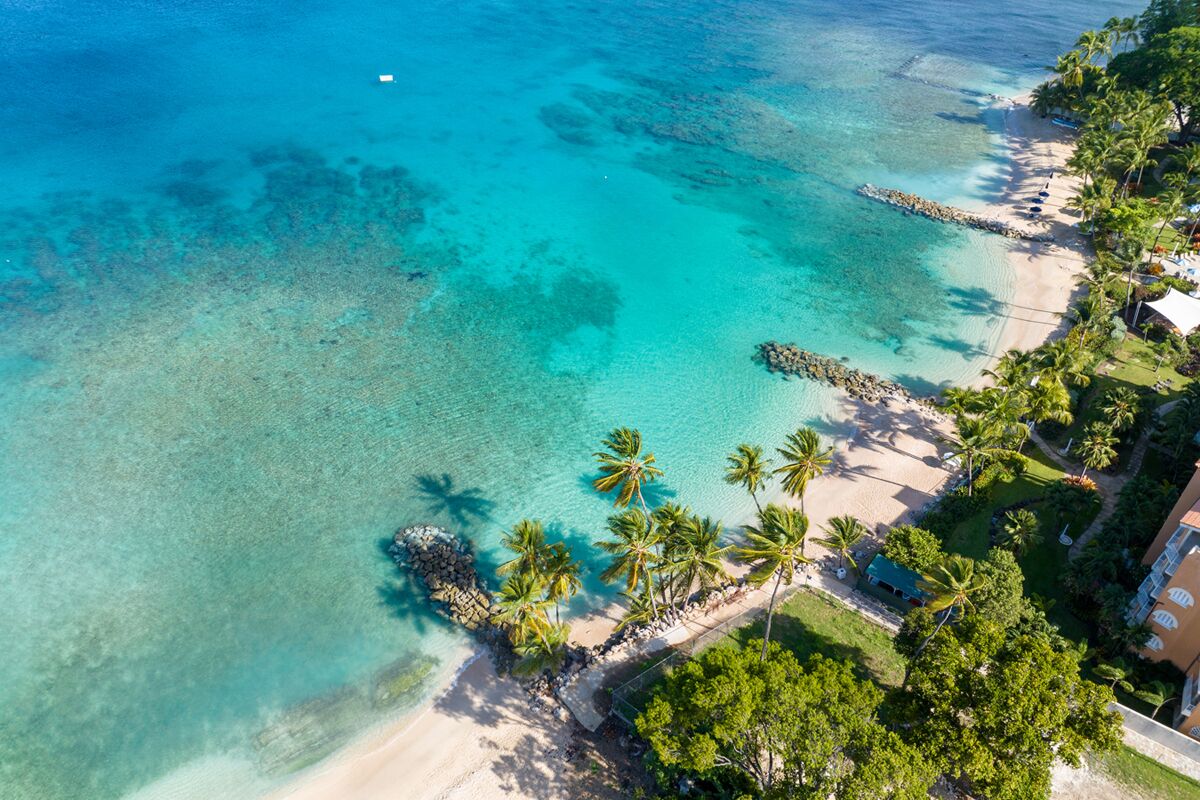 An aerial view of a beach with clear turquoise waters, palm trees, and a building with a balcony overlooking the beach.
