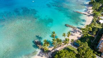 An aerial view of a beach with clear turquoise waters, palm trees, and a building with a balcony overlooking the beach.