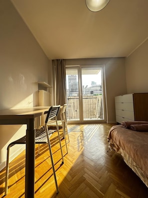 A room with a bed, a desk, and a chair, with a view of buildings through a large window. - Zlota 3-bedroom apartment in City Center,Pet Friendly (Warszawa)