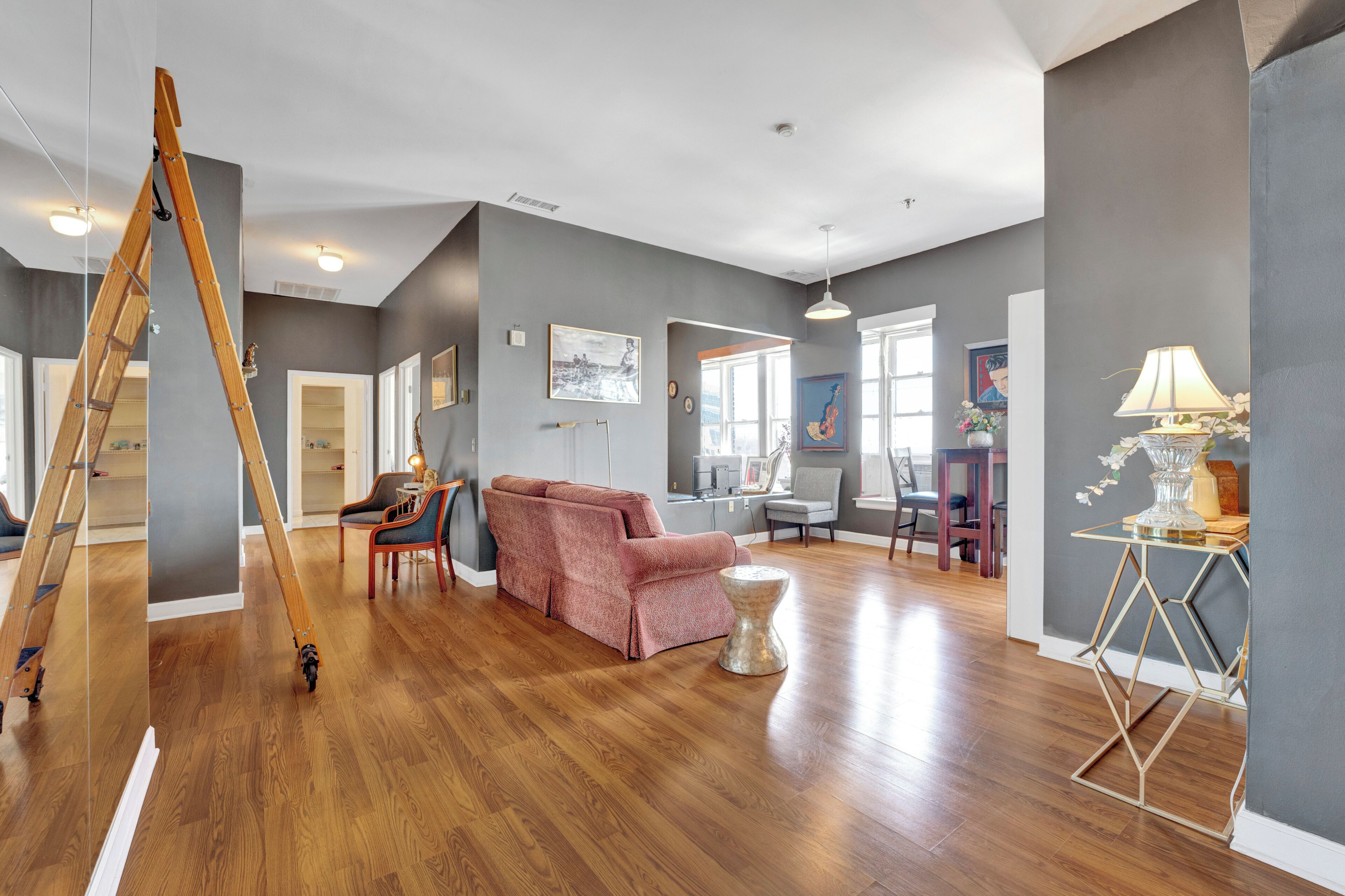 A spacious living room with hardwood flooring, a pink sofa, a wooden ladder, and a piano.