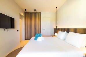 Ein Hotelzimmer mit Bett, einem an der Wand montierten Fernseher, einer Nachttischlampe und einer Tür zu einem weiteren Raum.