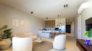 Un salon moderne avec un canapé, des fauteuils, une table basse et un coin repas, la cuisine étant visible au fond.