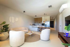 Ein modernes Wohnzimmer mit einer Couch, Sesseln, einem Couchtisch und einem Essbereich, im Hintergrund ist eine Küche zu sehen.