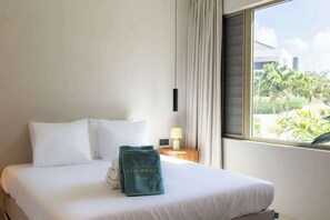 A hotel room with a bed, white linens, a teal towel, a nightstand with a lamp, and a window with a view of greenery. - Dolce Vita - Tropical Poolside Paradise (Willemstad)