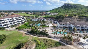 A resort with multiple buildings, a swimming pool area, and a view of a green hill. - Valen 1 | Elevated Caribbean Living at The Reef (Willemstad)