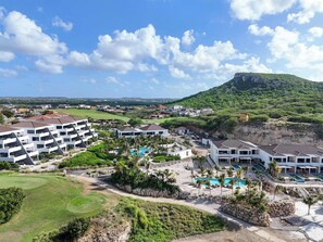 A resort with multiple buildings, a swimming pool area, and a view of a green hill. - Valen 1 | Elevated Caribbean Living at The Reef (Willemstad)