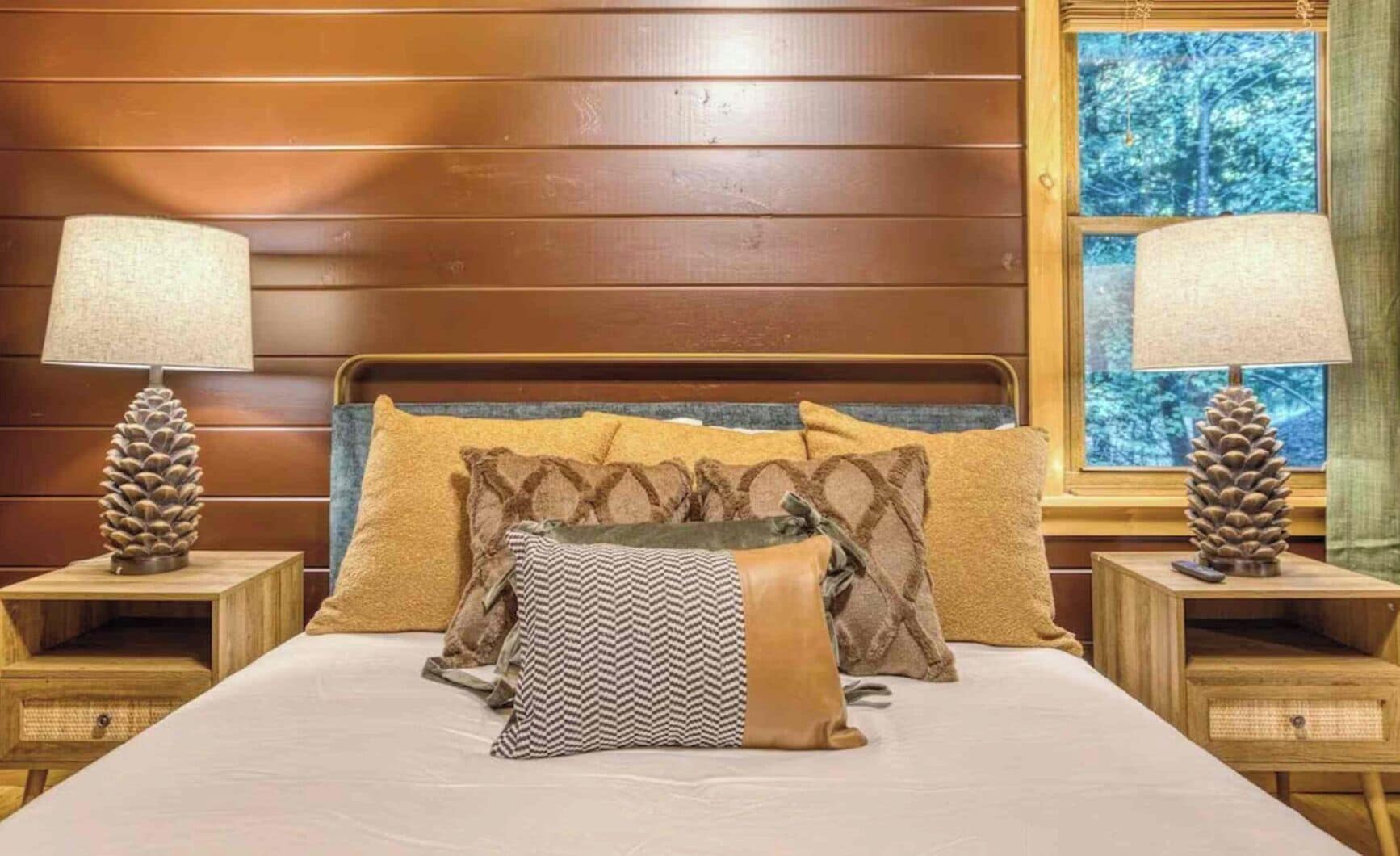 Une chambre à coucher avec une tête de lit en bois, un lit recouvert de plusieurs oreillers, deux tables de chevet ornées de lampes en forme de pomme de pin, et une fenêtre donnant sur la forêt.