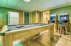A pool table with pool cues and balls, a refrigerator, and two arcade machines in a room with wooden walls and a tiled ceiling.