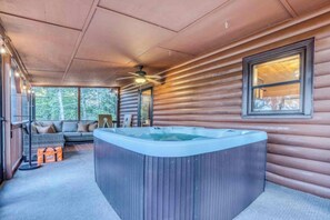 A hot tub in a cabin with a ceiling fan, a sofa, and a window overlooking a forest.