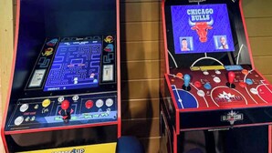 Zwei Arcade-Automaten, einer mit der Aufschrift 'PAC-MAN' und der andere 'NBA JAM', mit Joysticks und Bildschirmen, die Spieldarstellungen zeigen.