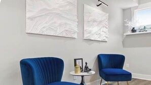 A room with two blue upholstered chairs, a small round table with a statue and a yellow container, and two abstract white wall art pieces.