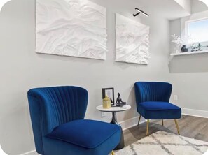 Una habitació amb dues cadires blaus amb tapisseria, una taula rodona petita amb una estàtua i un recipient groc, i dues peces d'art mural blanc abstracte.