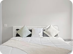 Una cama con cabecera blanca, varios almohadones y una colcha blanca.