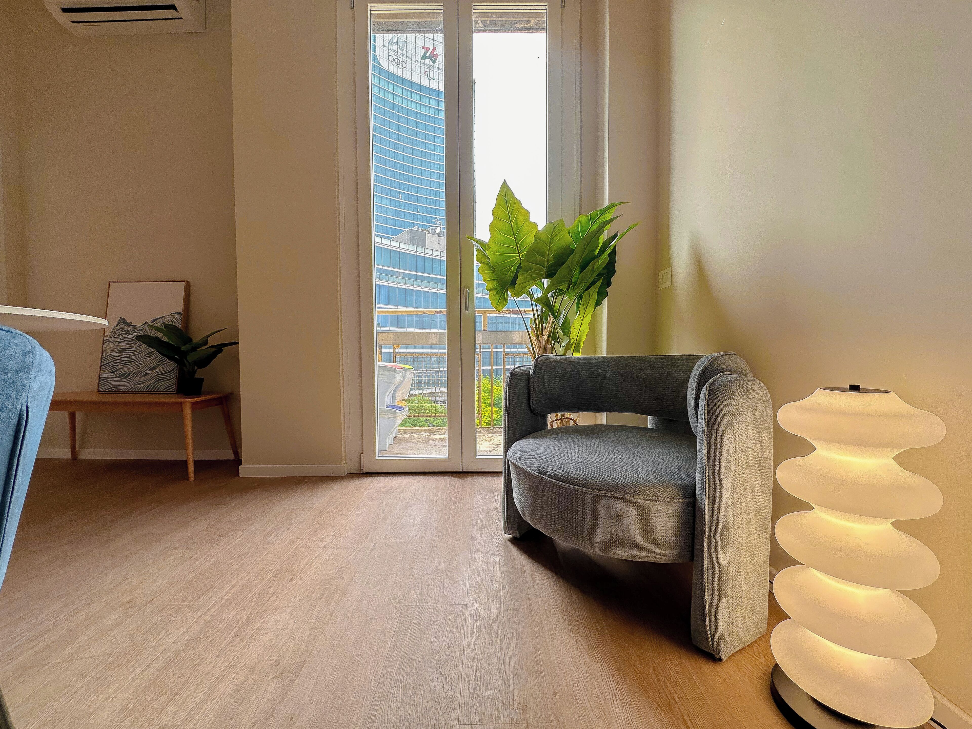 A modern room with a grey sofa, a potted plant, a lamp, and a view of a building through a glass door.