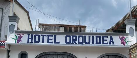 A hotel named 'HOTEL ORQUIDEA' with a white facade, black wrought iron window and door, and a small balcony with a flower decoration.