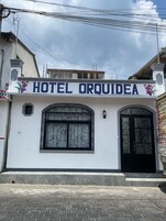 Hotel llamado 'HOTEL ORQUIDEA' con fachada blanca, ventanal y puerta de hierro forjado negro, y un pequeño balcón decorado con flores.