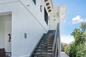 Un escalier menant à l’entrée d’un immeuble, avec une rampe, le numéro « 201 » au-dessus de la porte et un ciel dégagé en arrière-plan.