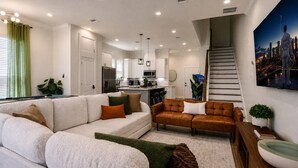 A modern living room with a sofa, a coffee table, and a staircase leading to an upper level. - Space City House | Hot Tub + Cinema | Sleep 16+ (Houston)