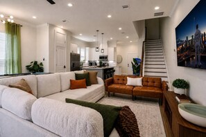 A modern living room with a sofa, a coffee table, and a staircase leading to an upper level. - Space City House | Hot Tub + Cinema | Sleep 16+ (Houston)
