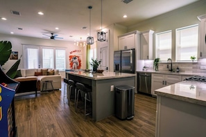 A modern kitchen with a central island, stainless steel appliances, and a dining area with a sofa and chairs. - Space City House | Hot Tub + Cinema | Sleep 16+ (Houston)