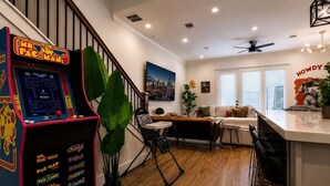 A living room with a retro arcade game, a staircase, a sofa, a coffee table, and a large plant. - Space City House | Hot Tub + Cinema | Sleep 16+ (Houston)