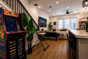 A living room with a retro arcade game, a staircase, a sofa, a coffee table, and a large plant. - Space City House | Hot Tub + Cinema | Sleep 16+ (Houston)