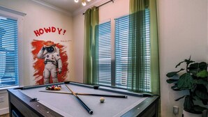 A pool table with a NASA astronaut mural, green curtains, and a potted plant. - Space City House | Hot Tub + Cinema | Sleep 16+ (Houston)