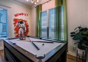 A pool table with a NASA astronaut mural, green curtains, and a potted plant. - Space City House | Hot Tub + Cinema | Sleep 16+ (Houston)