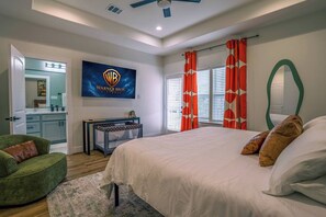 Ein Schlafzimmer mit einem großen Bett, einem Fernseher mit dem Warner Bros. Pictures Logo, einem Schreibtisch mit einem Computer und einem grünen Sessel.