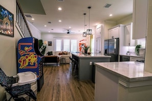 A modern kitchen with a central island, a dining area with a sofa, and a staircase with a decorative wall. - Space City House | Hot Tub + Cinema | Sleep 16+ (Houston)