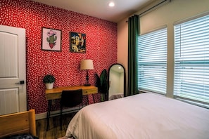 A bedroom with a polka dot wallpaper, a bed, a desk with a chair, a mirror, and a window with blinds. - Space City House | Hot Tub + Cinema | Sleep 16+ (Houston)