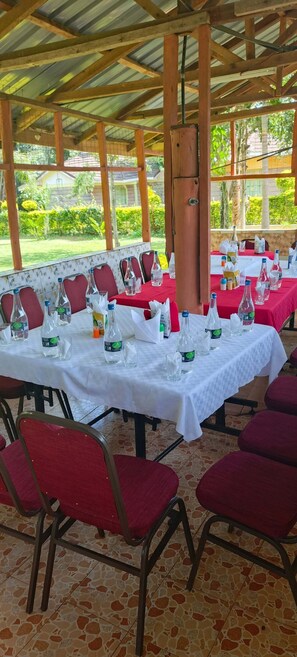 An outdoor covered dining area with tables set for a meal, chairs, and a view of the surrounding greenery.
