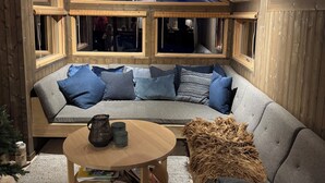Ein gemütlicher Raum mit Holzbalkendecke, einem grauen Sofa mit blauen Kissen, einem runden Holztisch mit einer Karaffe und Büchern sowie einem gewebten Pouf.